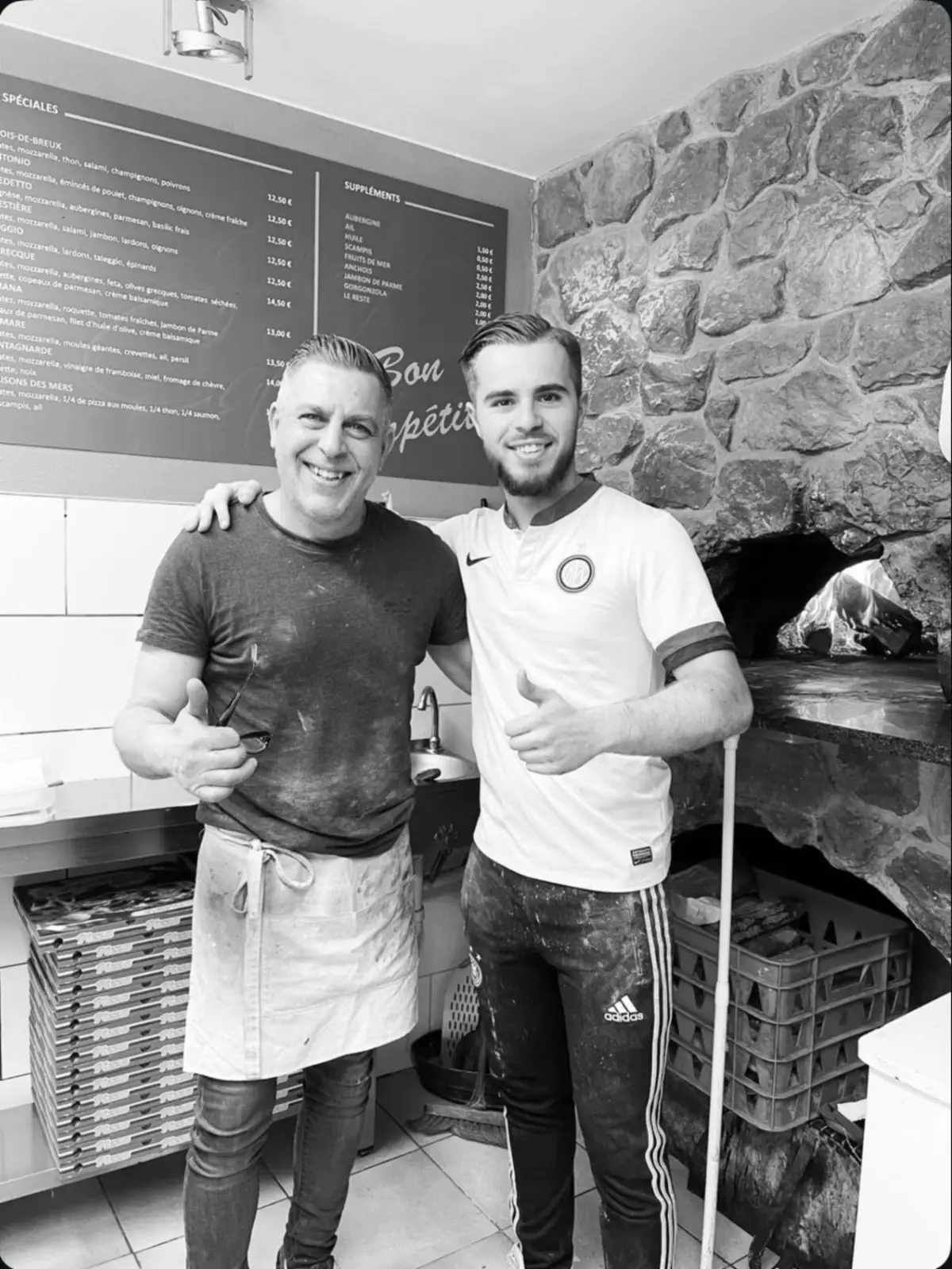 Les deux pizzaiolos de Chez Carlo devant le four à bois, le tableau de la carte derrière eux.