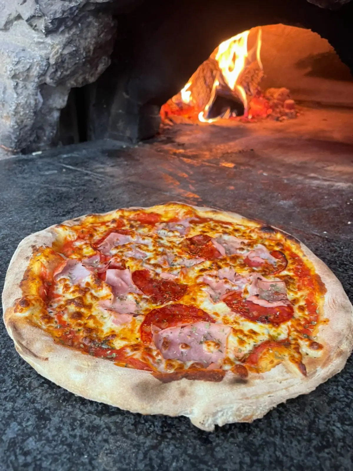 Une pizza qui finit de cuire sur la pierre du four à bois de Chez Carlo, la flamme visible au fond.