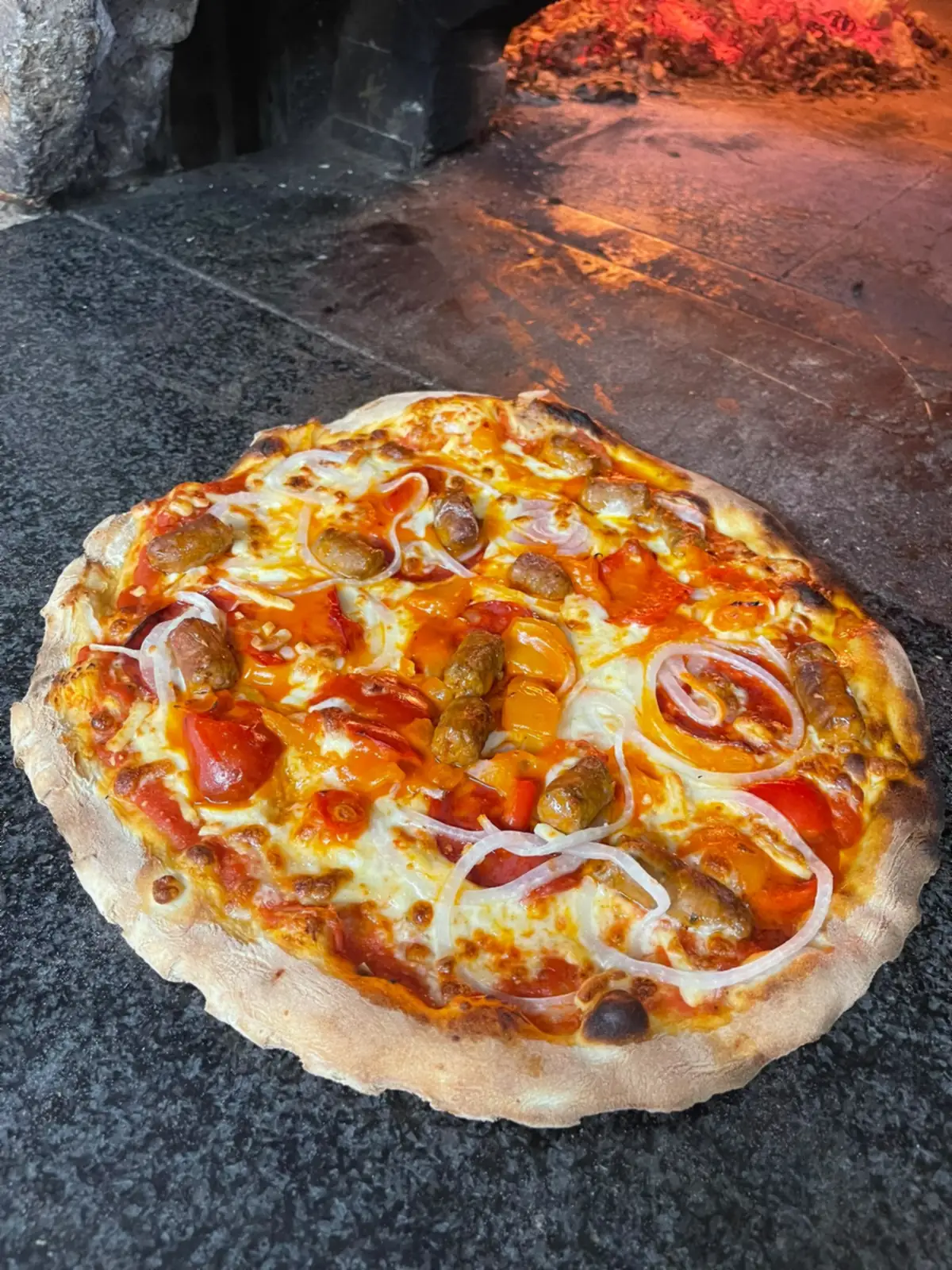 Une pizza aux légumes sur la sole du four, la flamme du bois derrière elle.